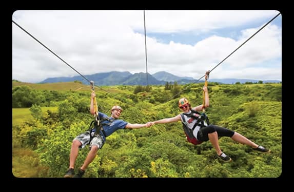  Two participants gliding on a dual zipline above green landscapes with scenic hills in the background