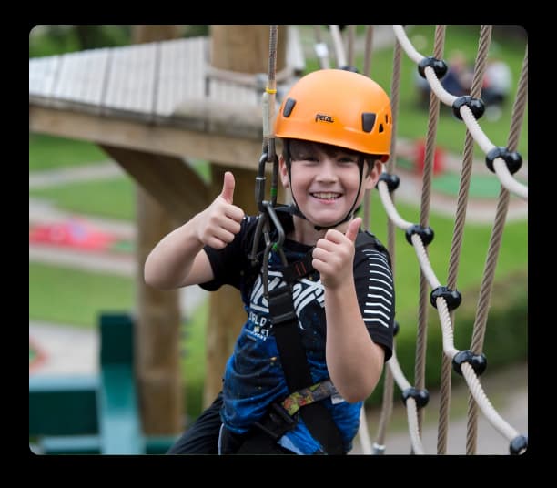 Smiling child wearing a helmet giving thumbs up while participating in a rope adventure course safely
