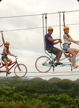 Participants on safety-harnessed bicycles ride on an elevated zip cycle trail with scenic views.